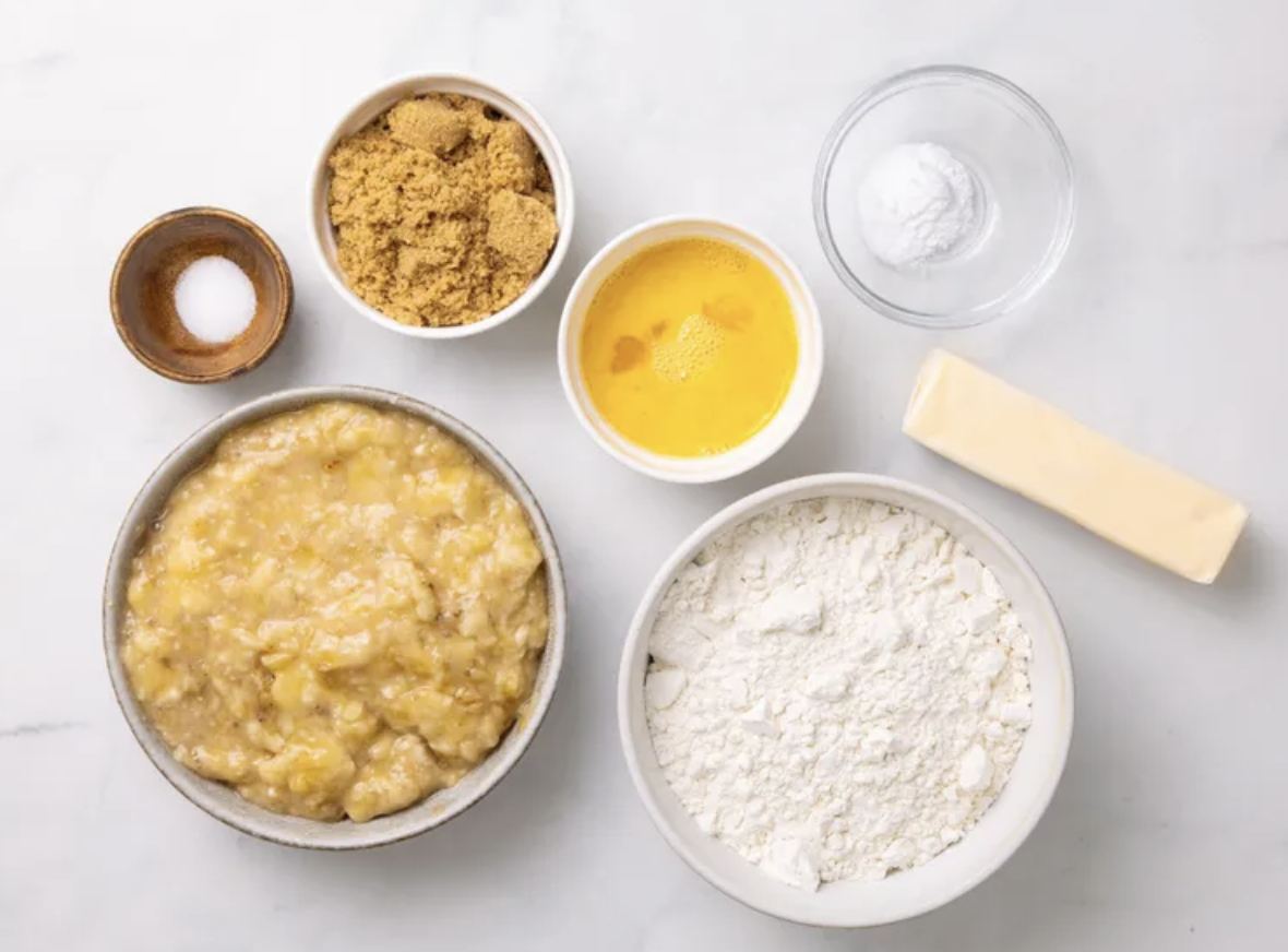 Bowls of mashed banana, brown sugar, flour, salt and baking soda on a marble counter