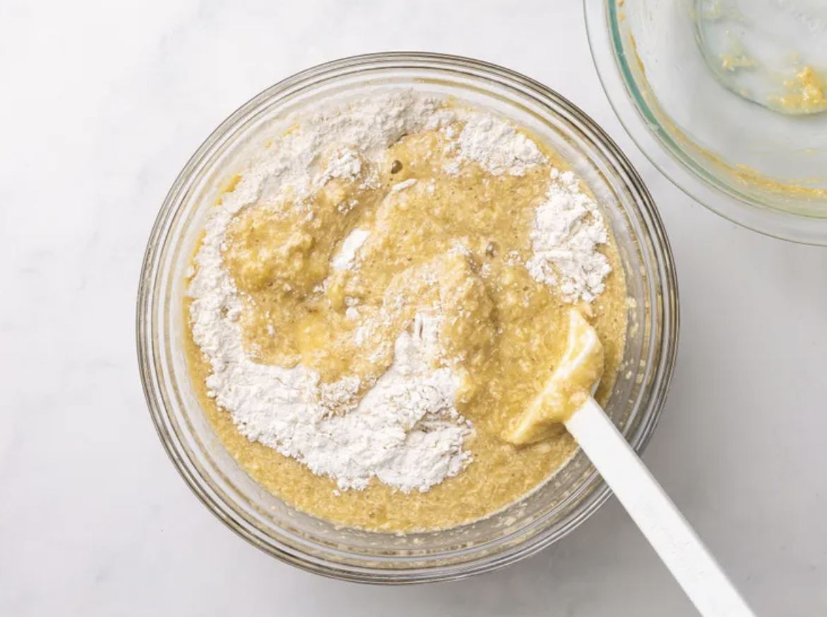 Flour being folded into banana batter in a glass bowl
