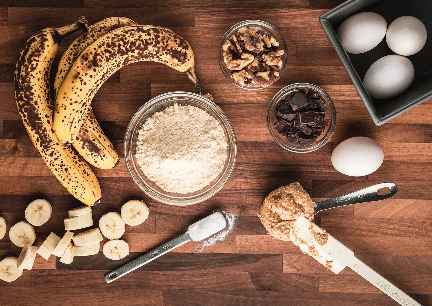 Overhead view of banana bread ingredients arranged on a wooden counter: ripe bananas, flour, walnuts, chocolate, eggs and measuring spoons