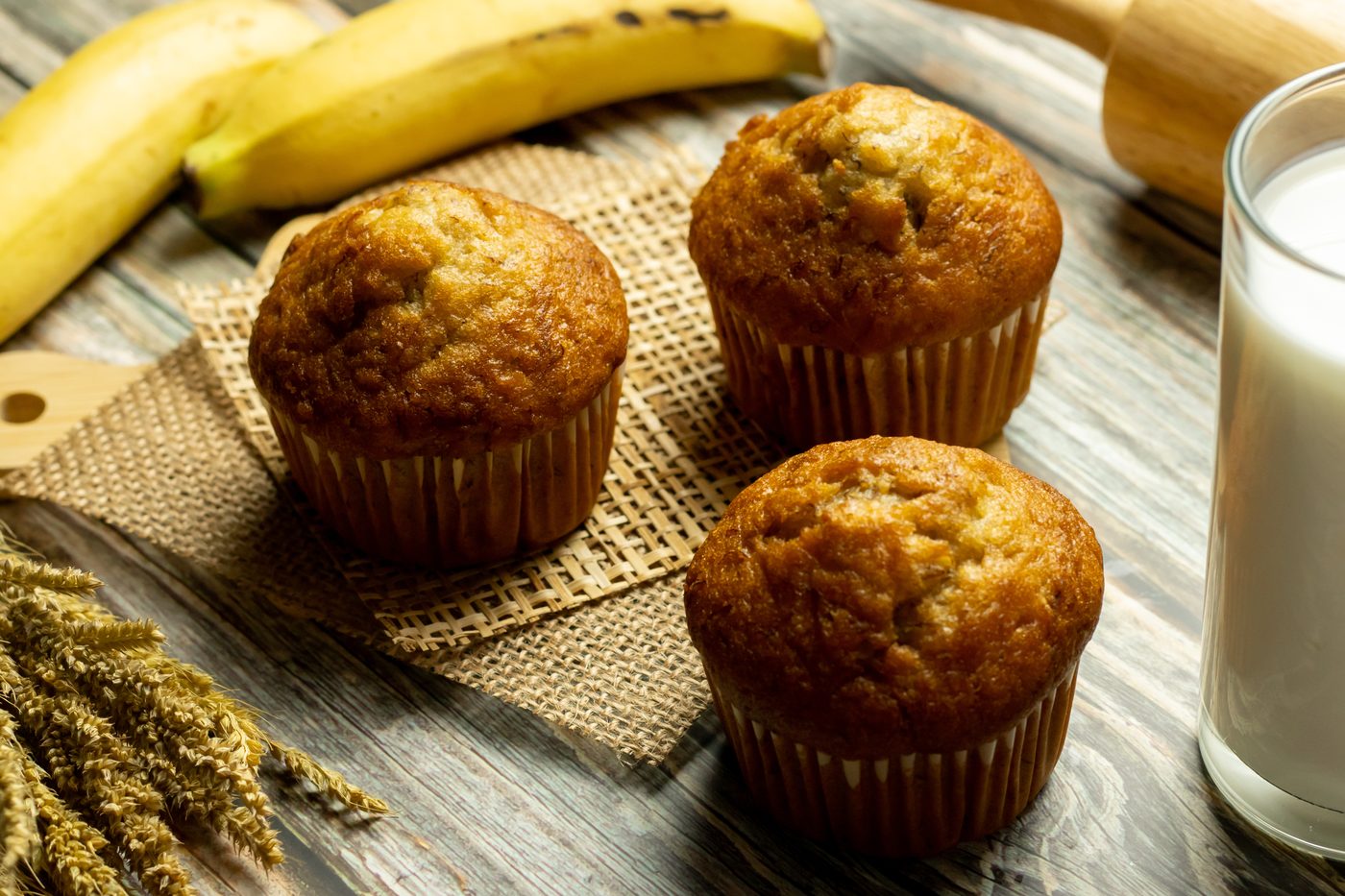Three banana muffins on burlap beside a banana and a glass of milk