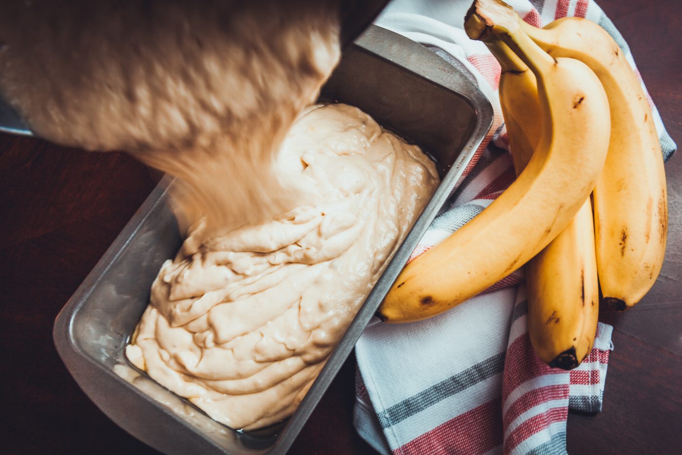 Batter being poured into a metal loaf pan, with bananas and a striped kitchen towel beside it